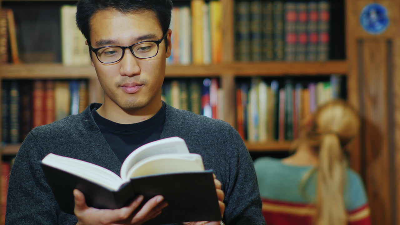hombre asiático guapo con gafas leyendo un libro en la biblioteca 1