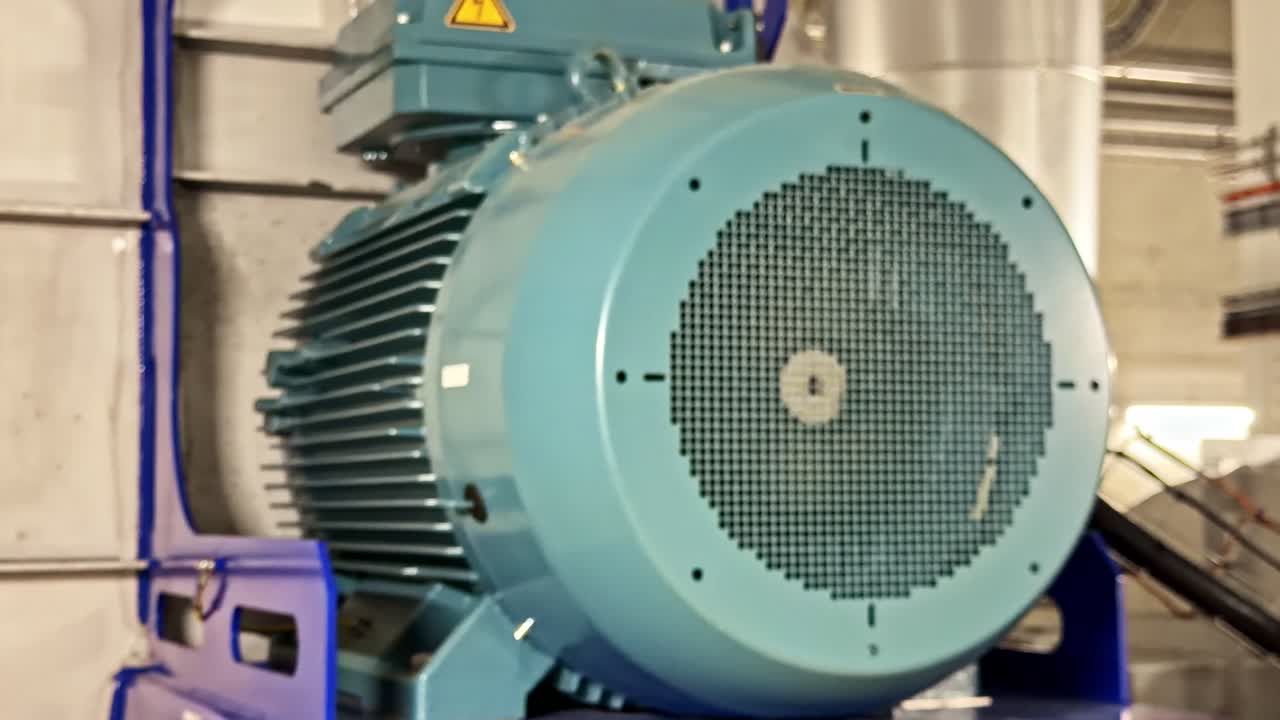 Primo piano di un motore elettrico blu funzionante con griglia di ventilazione ed etichetta di avvertenza montato in un impianto industriale circondato da tubature e macchinari