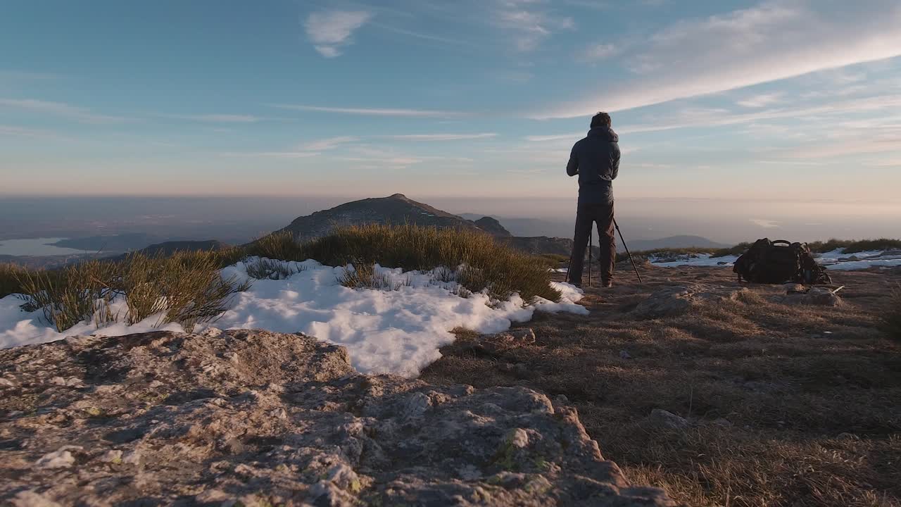 vista posterior de gran angular de un joven excursionista tomando fotos con su cámara y trípode en la cima de una montaña nevada en las montañas de guadarrama, madrid, españa