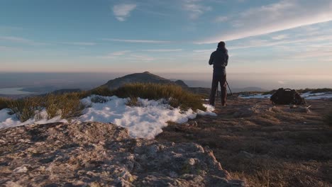 Vista-Posterior-De-Gran-Angular-De-Un-Joven-Excursionista-Tomando-Fotos-Con-Su-Cámara-Y-Trípode-En-La-Cima-De-Una-Montaña-Nevada-En-Las-Montañas-De-Guadarrama,-Madrid,-España