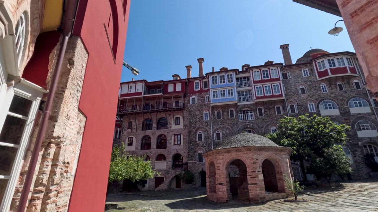 Visitors walk through a historical site at Mount Athos in Greece. They admire the buildings and take in the beauty of the surroundings. The sun shines bright in the clear sky.