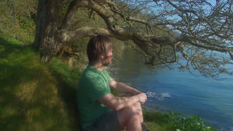 alone man relaxing while leaning on grassy hill under the tree at seaside. - close up