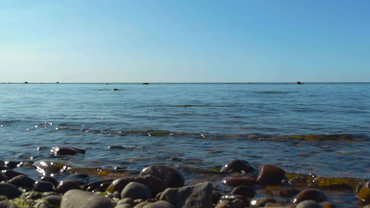 Low angle slow motion footage of a rock or a pebble being thrown and is flying by the footage and skipping on the blue ocean sea water during a summer sunny day. mossy and glossy rocks in the front