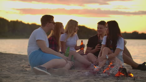 the best friends from university are sitting in around bonfire on the lake coast. they are talking to each other and drinking beer at sunset.