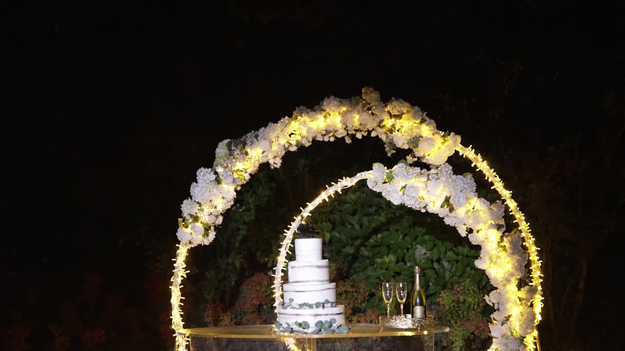 Wedding cake displayed under a floral arch with soft lighting and champagne glasses nearby