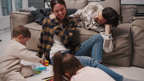 A-happy-brunette-girl-in-a-plaid-shirt-and-blue-jeans-sits-on-the-floor-and-plays-with-her-children-her-husband-lies-near-them-on-the-sofa-they-spend-fun-time-together-in-a-modern-studio-apartment