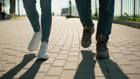 close-up of legs in motion, individuals walking closely on an interlocked pathway wearing sneakers and boots under bright sunlight with blur background featuring iron railing