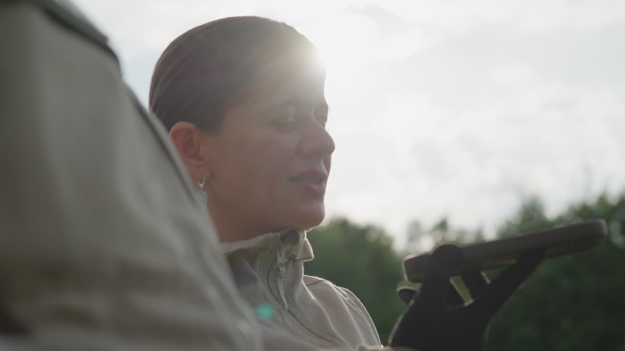 close up of woman shading eyes with gloved hand while holding mobile device, sunlight flare through clouds creating lens glow, blurred colleague shoulder and tree line backdrop
