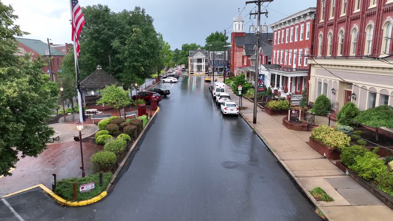 Low angle flight over wet rainy street with cars in small american town. Flag of USA in historic town. Church and buildings during rainy day.