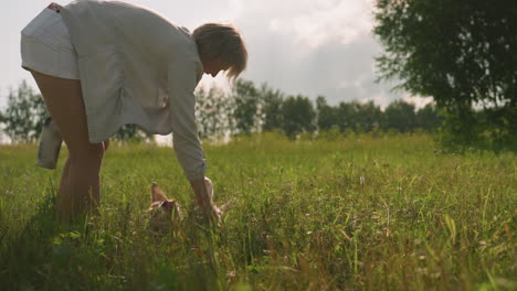 woman with left hand holding something while playing with her dog as dog happily rolls on grassy field under sunny sky, bright sunlight shines over open landscape