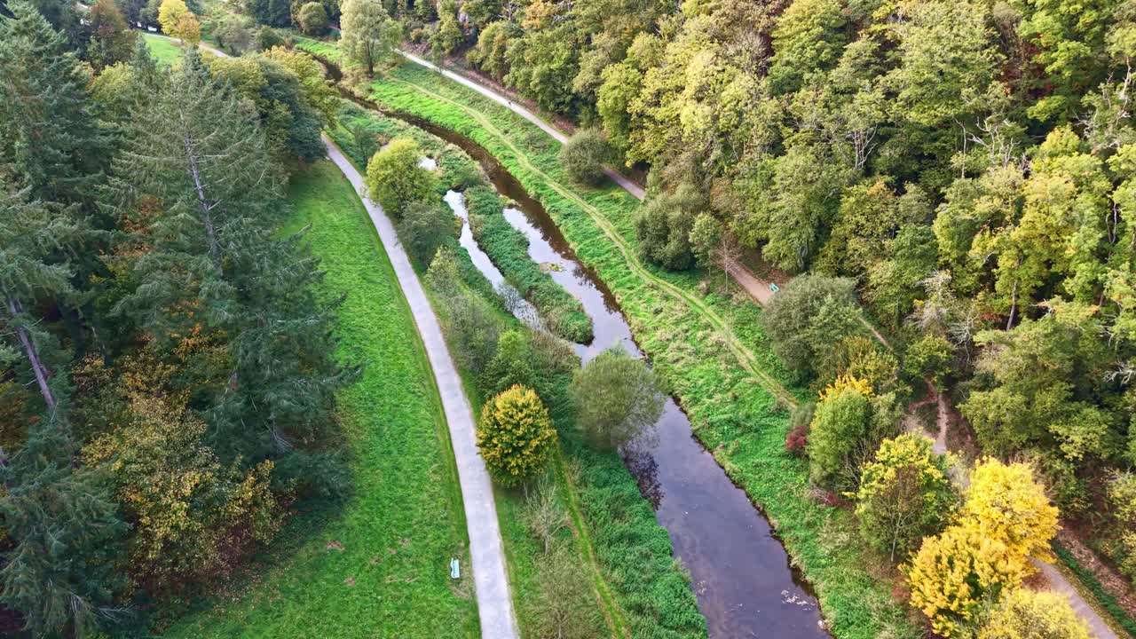 Tilted drone fly at winding water channel flowing through on green Concise Forest in autumn scenery, Saint-Berthevin, Mayenne, France