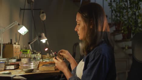 focused caucasian female jeweller sitting at desk, making jewelry in workshop