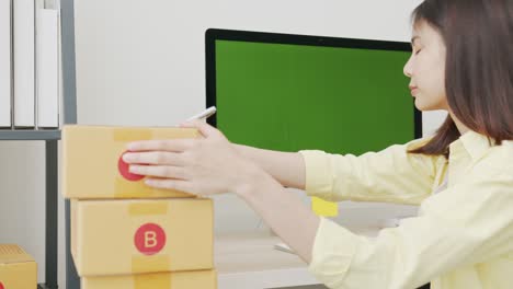 startup small business, young asian woman checking online order on computer with green screen. and packing boxes for products to send to customers.