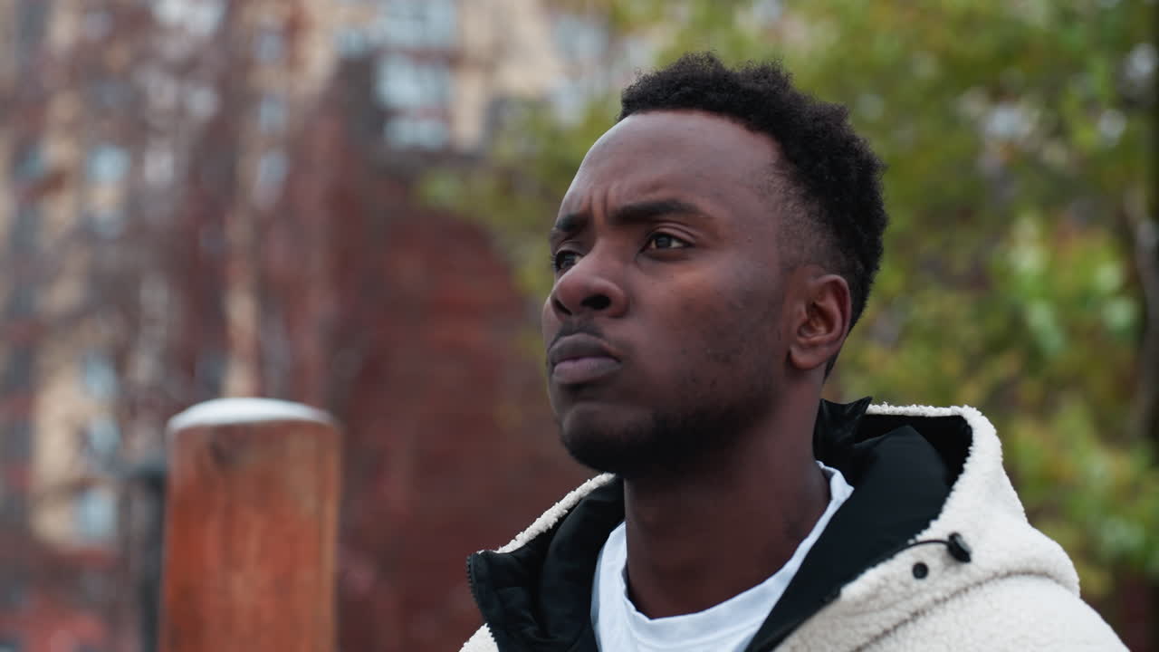 Rap artist in winter jacket looking focused while blinking eyes, wooden structure and blurred urban background with trees create a moody and intense atmosphere