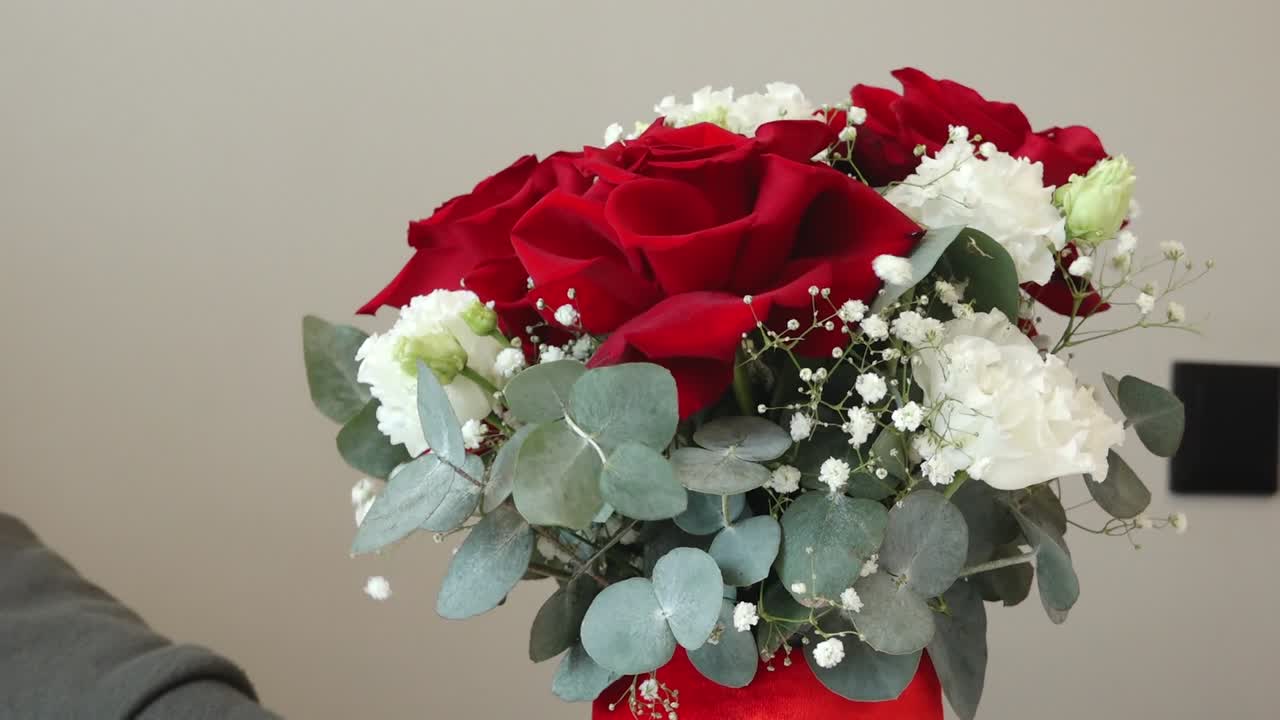 a person holding a bouquet with a vibrant red rose, delicate white baby's breath, and eucalyptus leaves. Soft indoor background emphasizes romance and elegance