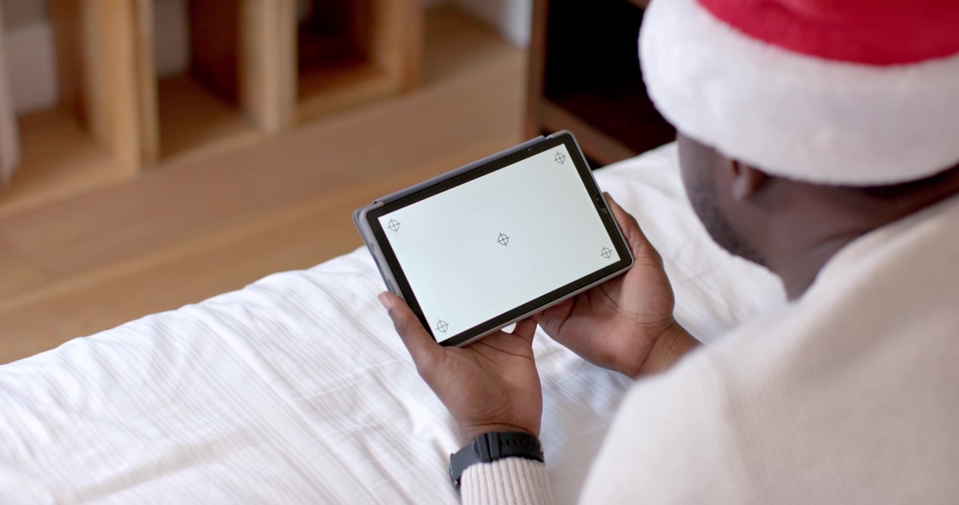 Man wearing Santa hat is adjusting grip and tilting tablet on bed to maintain screen focus