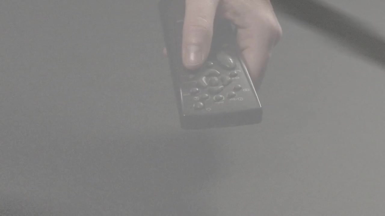 Hånd kommer ind fra toppen, griber fjernbetjening, tommelfinger glider på styreflade, slipper efter at have tjekket kontroller.