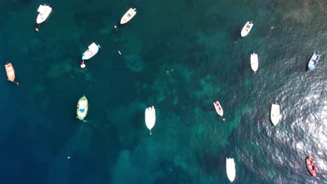 Vista-De-Arriba-Hacia-Abajo-De-Los-Barcos-De-Pesca-En-El-Mar-Mediterráneo-Con-Agua-Azul-Cristalina-Revelando-En-Santorini,-Grecia