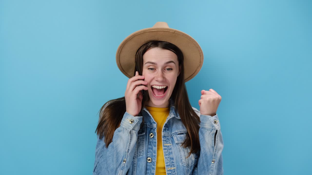 feliz joven emocional hablando por teléfono hace el gesto de sí levanta el puño cerrado con alegría recibe noticias impresionantes, usa chaqueta de denim y sombrero, aislado en fondo azul. concepto de estilo de vida de la gente