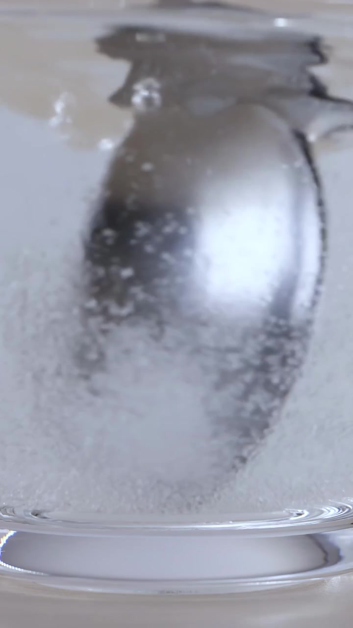 Primo piano di un cucchiaio di metallo che mescola lo zucchero nell'acqua calda all'interno di un bicchiere trasparente