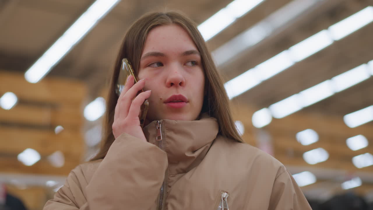Close-up of a young girl wearing a light brown jacket, standing in a mall, holding a phone to her ear as she engages in a conversation with a slight smile