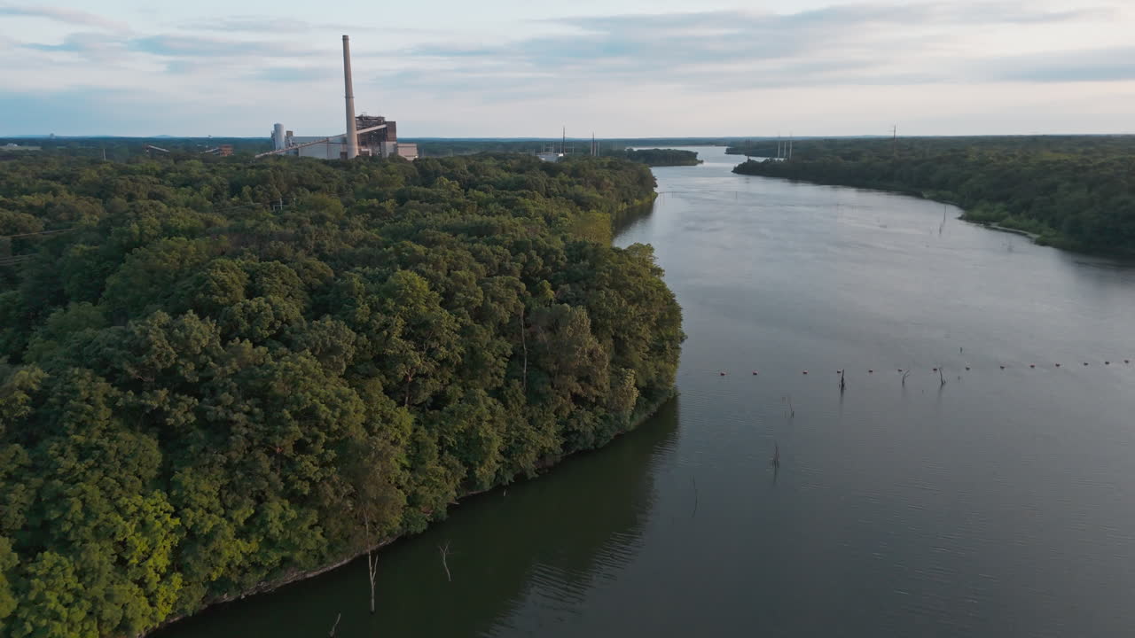 Drone-tilbagetrækning langs bred søoverflade, der strækker sig mod skovklædt kystlinje med bløde, blege refleksioner, Flint Creeks kraftværk i horisonten