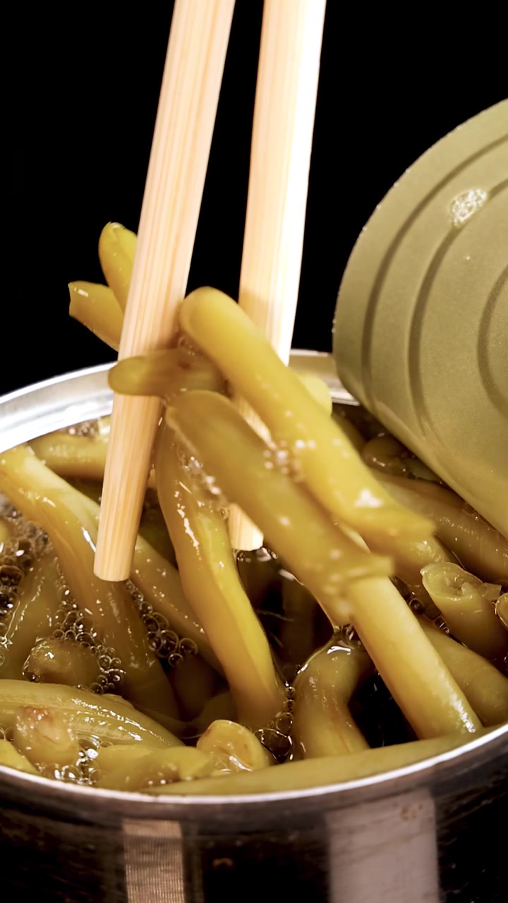 Wooden chopsticks grasp and lift haricot beans from an open can of canned food, under bright studio lighting with a black background and close-up framing