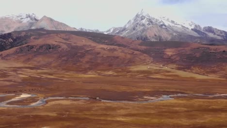 aerial drone footage panning from right to left revealins dark brown tibetan plateau with large white snow capped mountains and a small river flowing in front of them in a desolate and rural land.