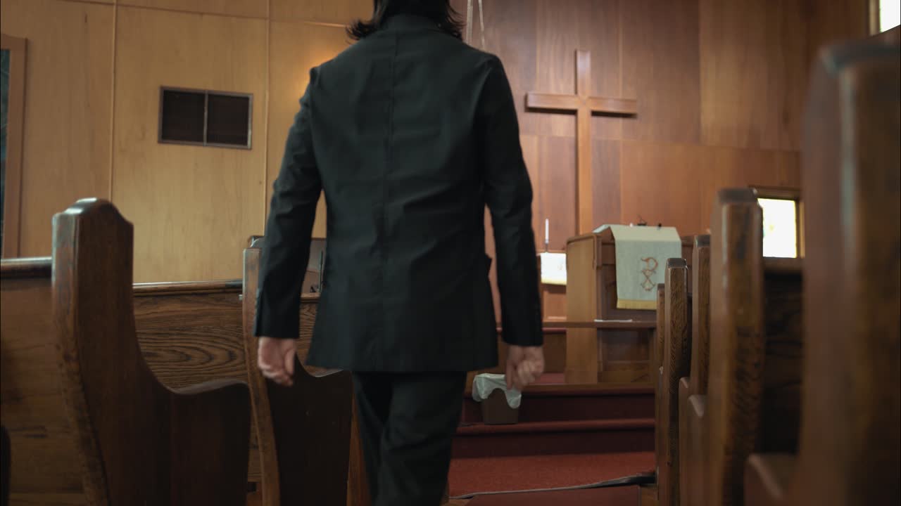 검은색 의상을 입은 목사가 교회에서 고 영화 속도 느린 동작으로 벤치에 앉아 있습니다.