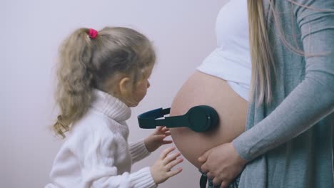 cute girl puts headphones on pregnant mommy belly in room