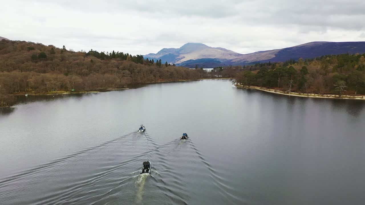 Filmisk luftbillede af en overflyvning af 3 både, der nærmer sig snævringen på Loch Lomond i Skotland, stiger op for at afsløre Ben Lomond.