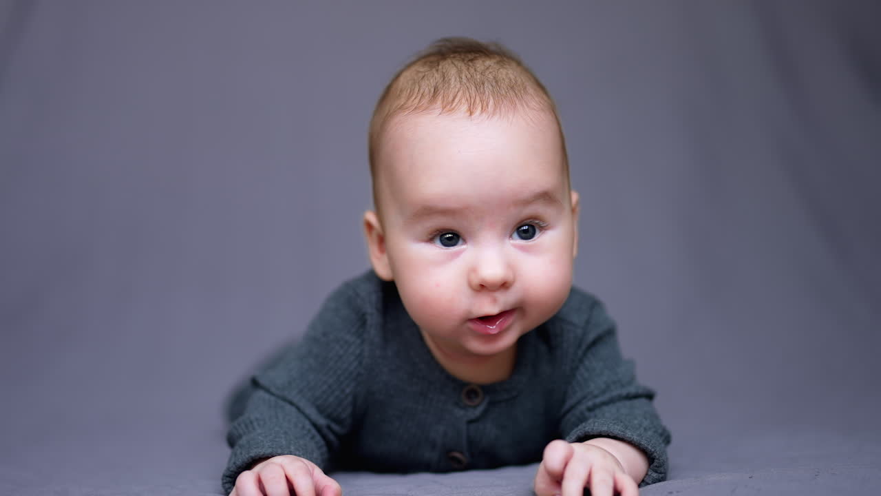 Adorable baby boy lies on bed and looks into camera. Beautiful child smiling sweetly. Grey backdrop.