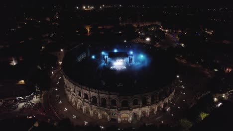 Drone-to-move-back-on-the-Arenas-de-Nîmes-in-the-middle-of-the-night,-people-are-watching-the-concert-and-there-are-lights-of-several-colors