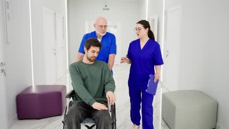 A-confident-elderly-doctor-in-glasses-with-a-gray-beard-and-in-a-blue-uniform-takes-a-man-to-wheelchairs-for-the-disabled-while-a-brunette-girl-talks-with-the-doctor-about-the-rehabilitation-of-a-young-brunette-guy-in-a-green-shirt-after-a-broken-leg