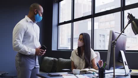 Diverse-business-people-wearing-face-masks-using-computer-talking-in-office