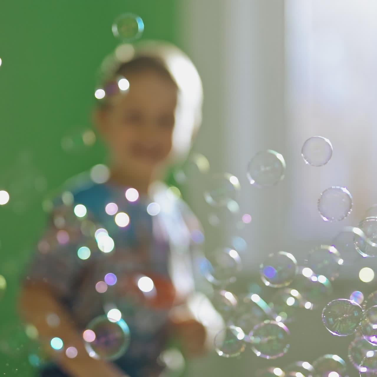 Round bubbles are flying inside the room and a boy catching and bursting them. Happy kid is playing with colorful soap bubbles at home.
