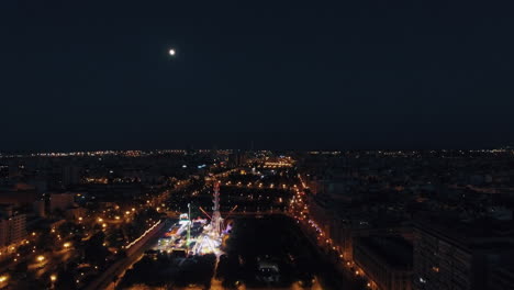 Aerial-night-view-of-lighted-ferris-wheel-in-amusement-park-against-sky-with-moon-Valencia-Spain