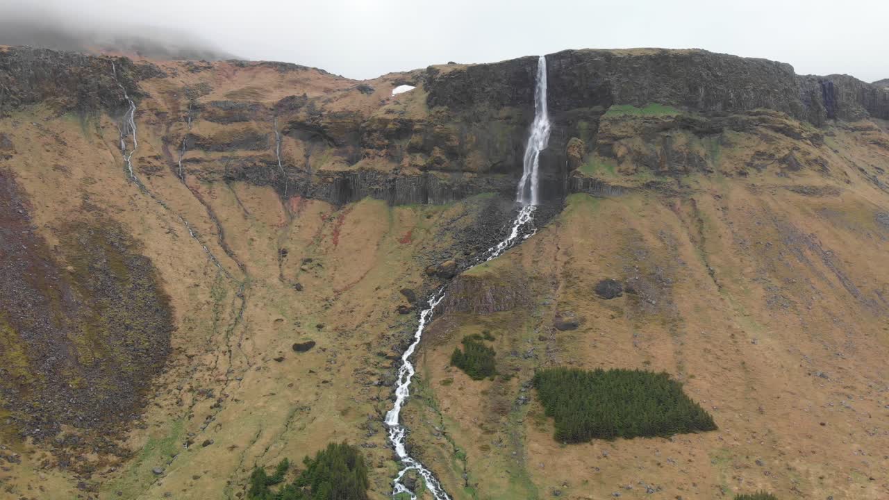 Aerial video of the magnificant mountains surrounding the tall and slim waterfall Bjarnarfoss that leads to a river in the beautiful Icelandic landscape on a misty and cloudy day