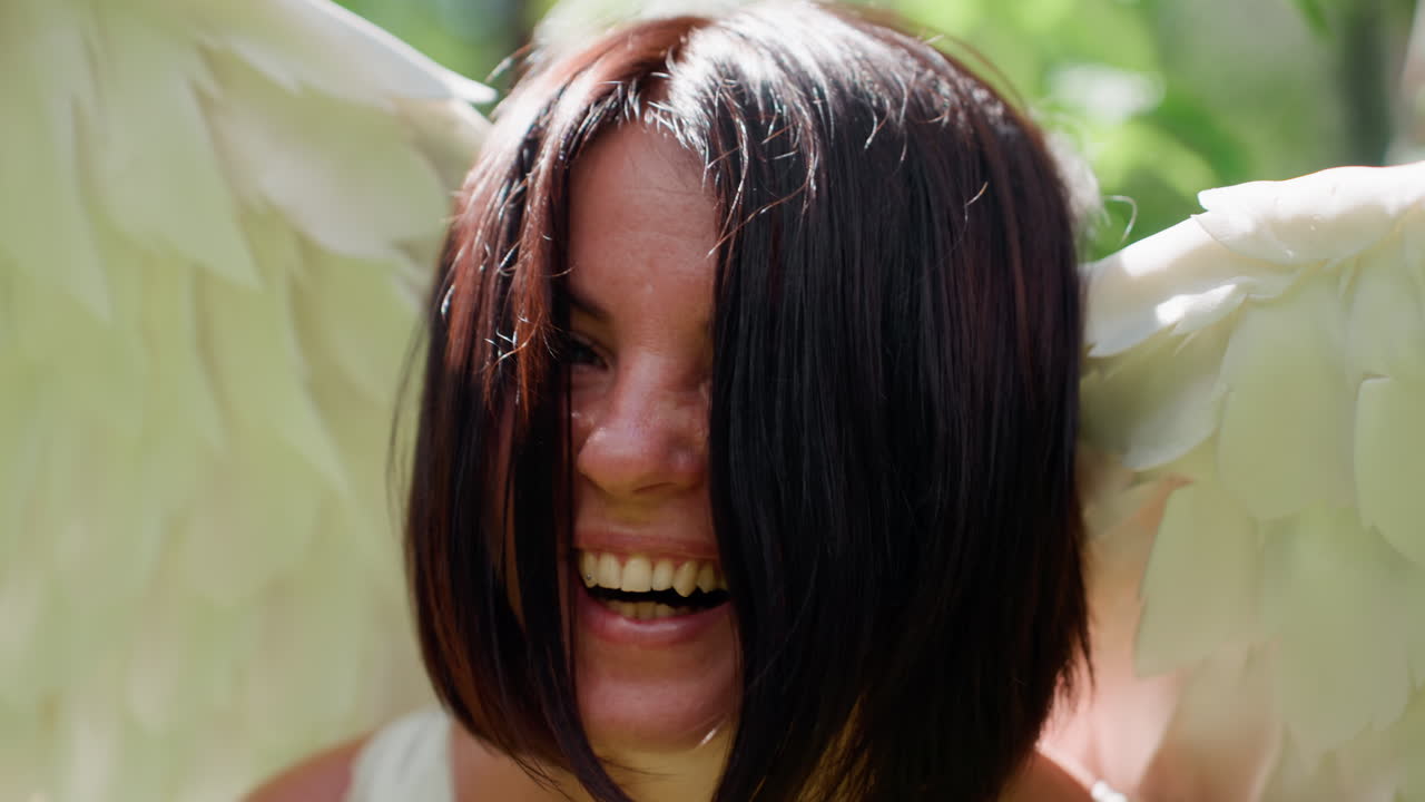 Close up of smiling woman with dark hair wearing soft white angel wings outdoors under sunlight, joyful expression radiating warmth and purity surrounded by bright ethereal glow