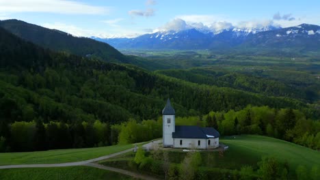 Iglesia-Con-Una-Fachada-Blanca-Y-Un-Campanario-Puntiagudo,-Rodeada-De-Exuberante-Vegetación,-Frente-A-Un-Cielo-Parcialmente-Nublado-Con-Montañas-Al-Fondo