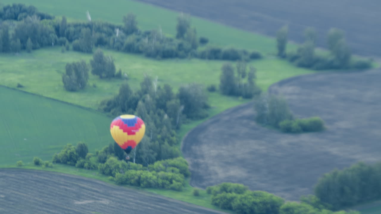Distant view of vibrant hot air balloon drifting above patchwork of green and brown fields dotted with small tree clusters, wicker basket suspended beneath colourful canopy in gentle light