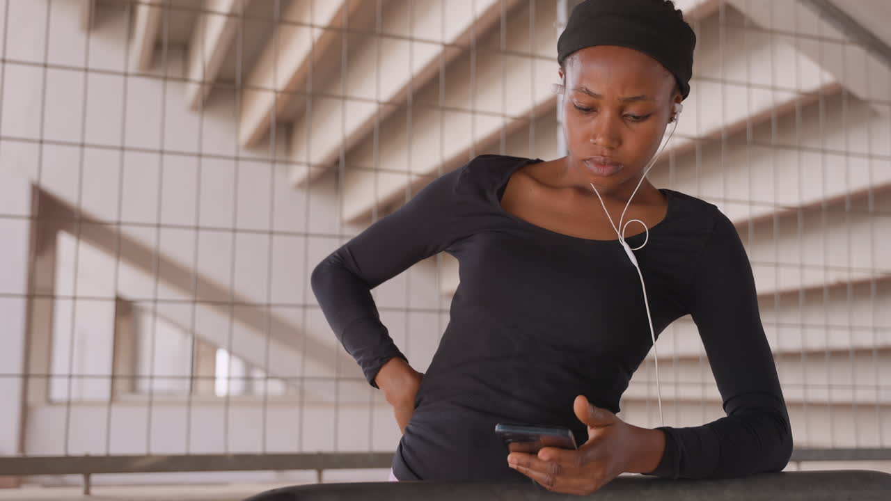 donna sulla ringhiera che controlla lo schermo del telefono. mani sui fianchi, auricolari pendenti, cavalcavia moderno, travi del soffitto ripetute, espressione concentrata, scansione dell'orario, pendolarismo urbano, toni neutri