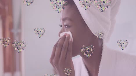 animation of floral hearts over happy african american woman cleansing face in bathroom