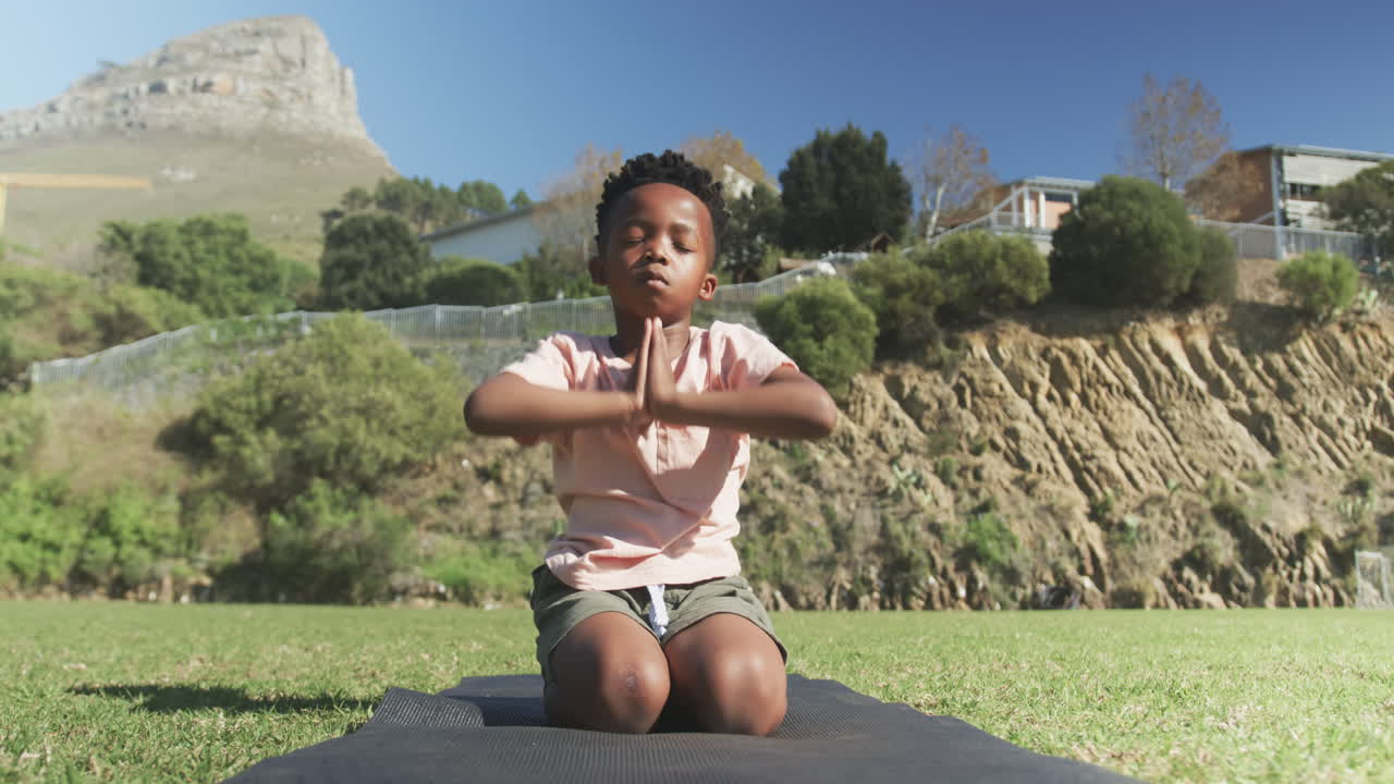 Meditating on yoga mat, African American boy practicing mindfulness outdoors in school setting