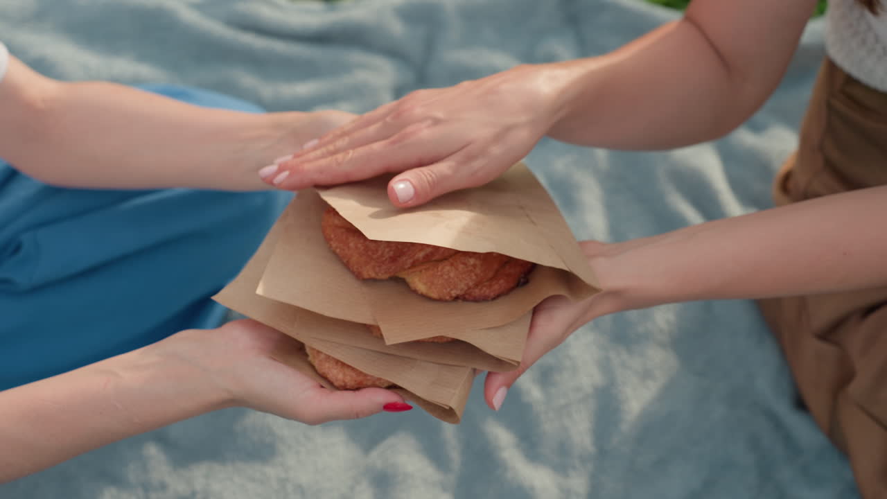 woman handing snacks to friend on picnic blanket in park, close hands exchange of wrapped pastries, warm sunlight and soft blurred grass background conveying friendly sharing moment
