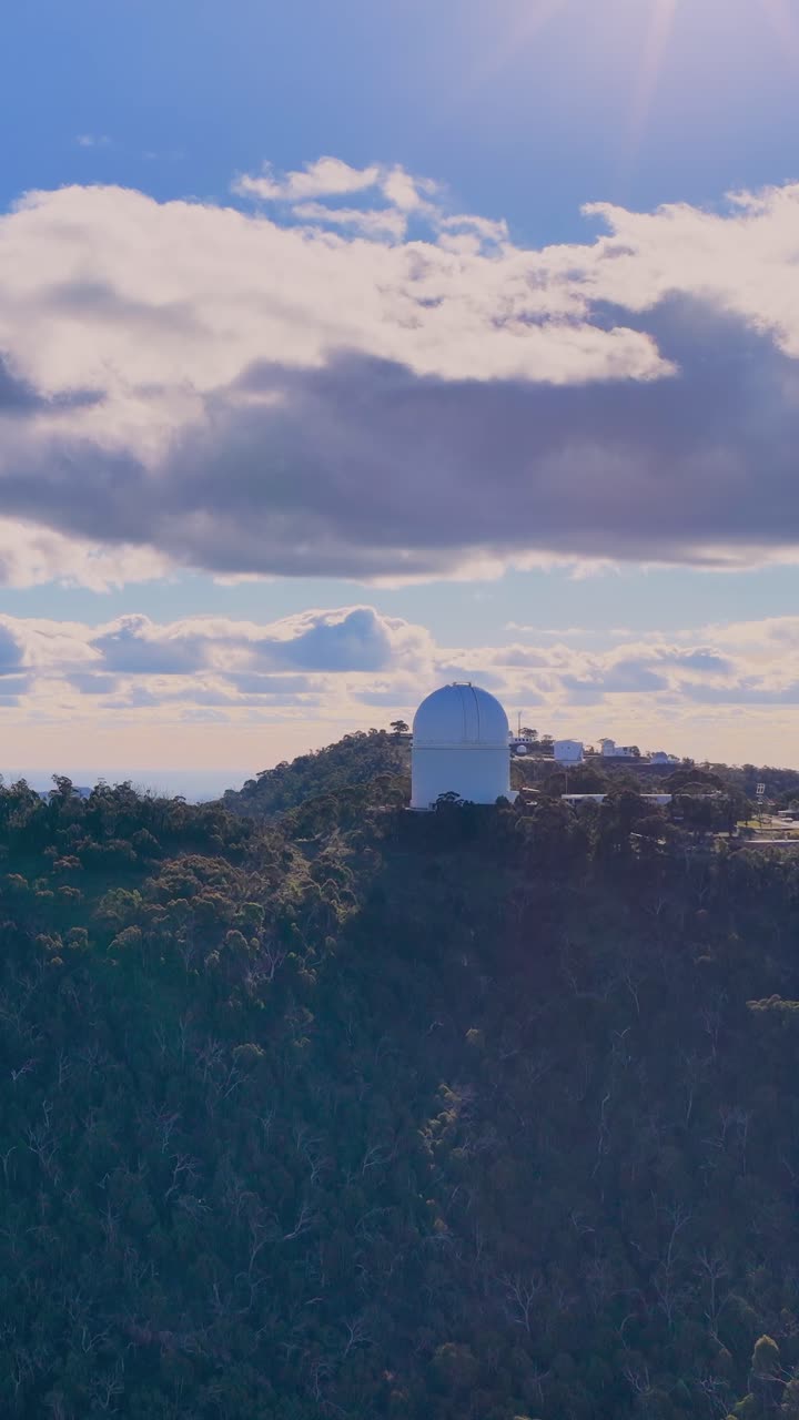 Dronekamera panorerer jævnt hen over en skovklædt bjergryg, afslører en stor hvid observatoriekuppel under dramatiske morgenskyer og blødt sollys.