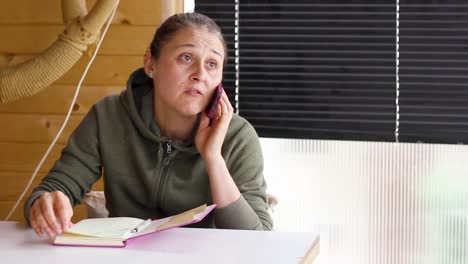 Woman-having-phone-conversation-sitting-at-table-looking-sideways,-panning-shot