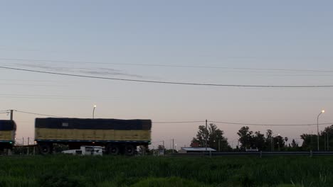 Un-Camión-Amarillo-Que-Transporta-Un-Remolque-Se-Mueve-De-Derecha-A-Izquierda,-Con-Cables,-Postes-Y-Un-Par-De-Casas-Al-Fondo-Al-Atardecer