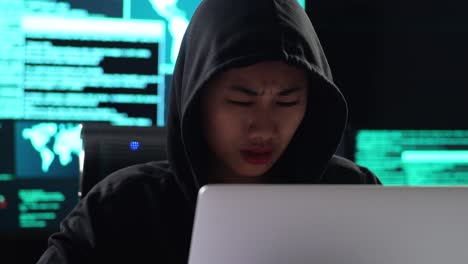 close up of asian female hacker in a hood having a headache while works on a laptop with maps and data on display screens of multiple monitors in a dark office room