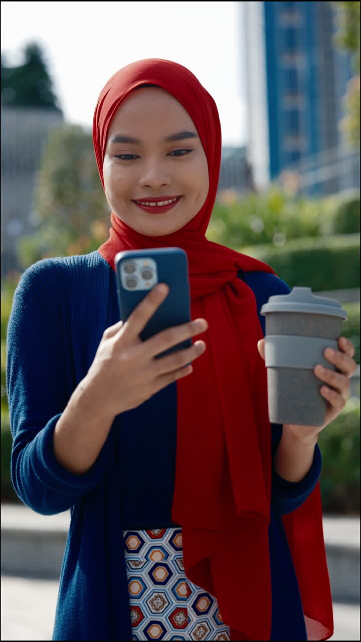 Young woman in hijab holding phone and reusable cup outdoors
